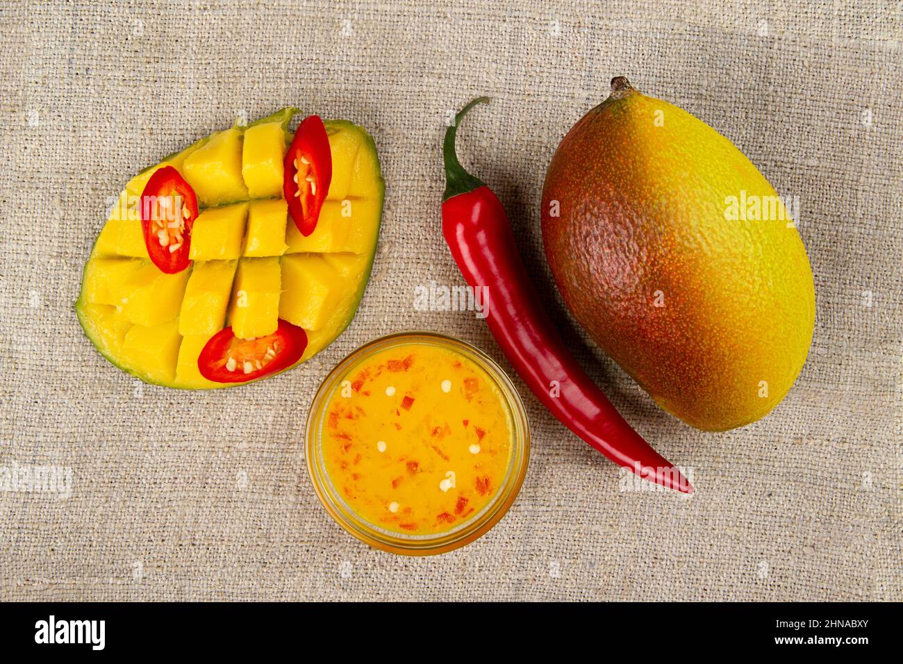 Top view of Whole mango, whole chili pepper, sliced hot pepper, cutted half of mango and Spicy mango chutney in glass bowl on burlap. Stock Photo