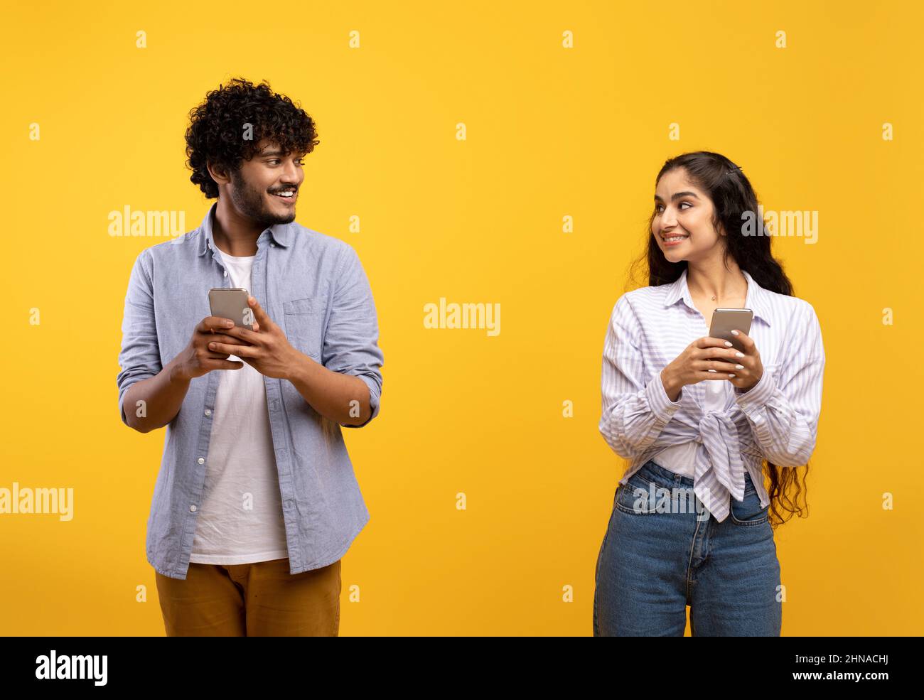Cheerful young indian man and woman in casual outfits holding smartphones, looking at each other and smiling Stock Photo