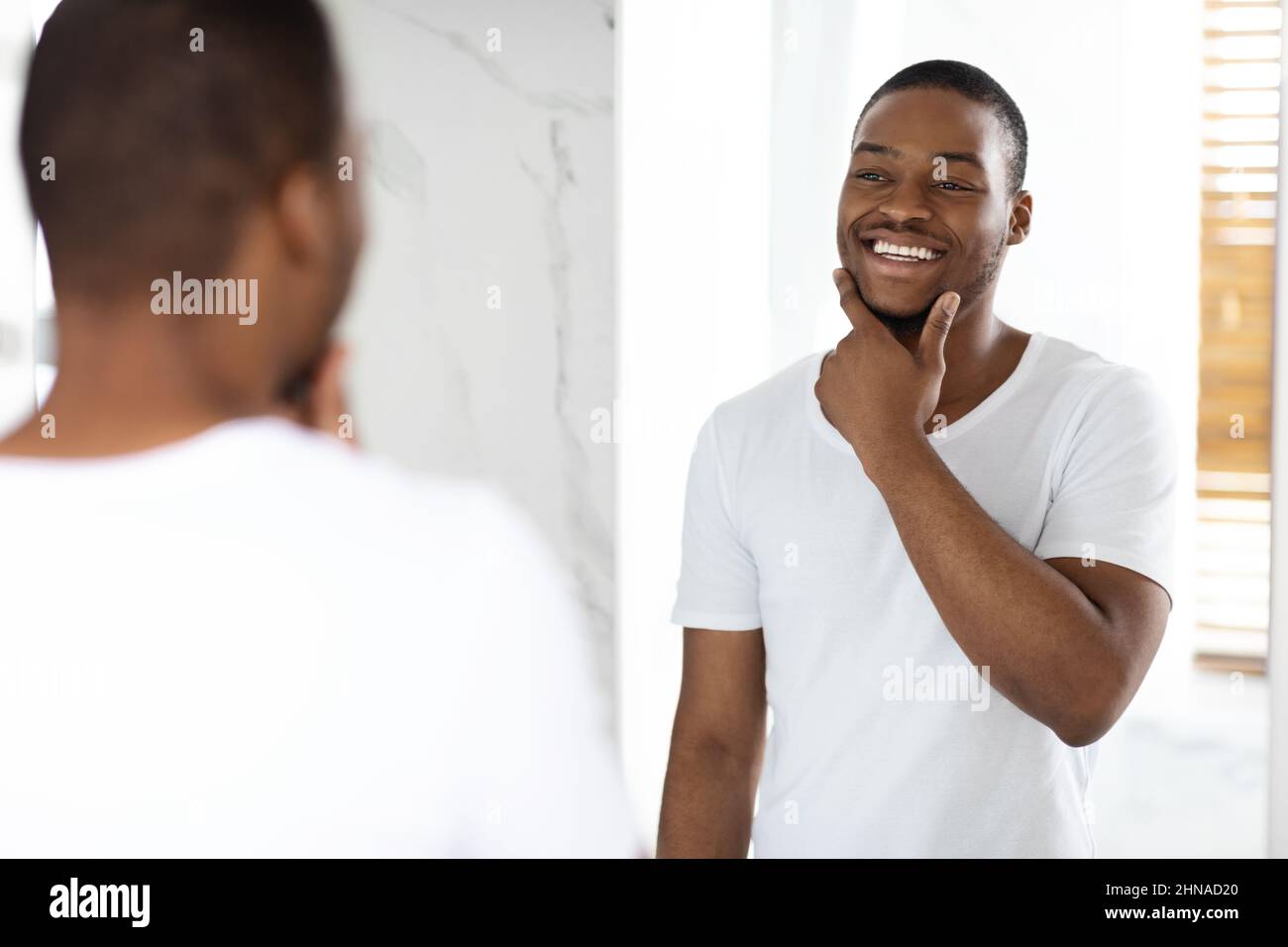 Happy Young Black Man Looking In Mirror In Bathroom And Touching Face