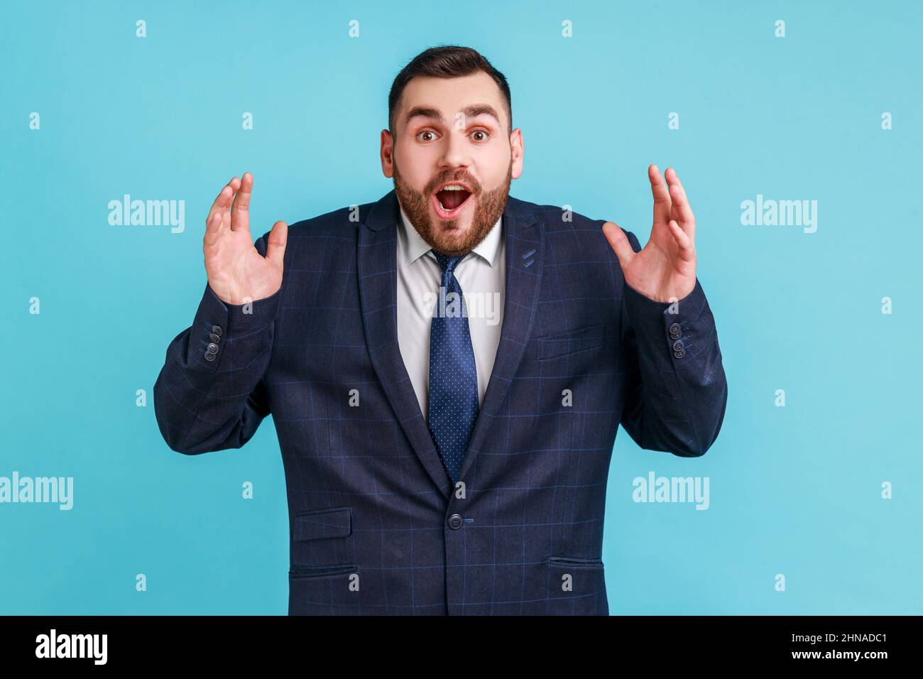 Wow, it's unbelievable. Surprised bearded man in official style suit staring at camera with widely open mouth and big eyes, raising arms in amazement. Indoor studio shot isolated on blue background. Stock Photo
