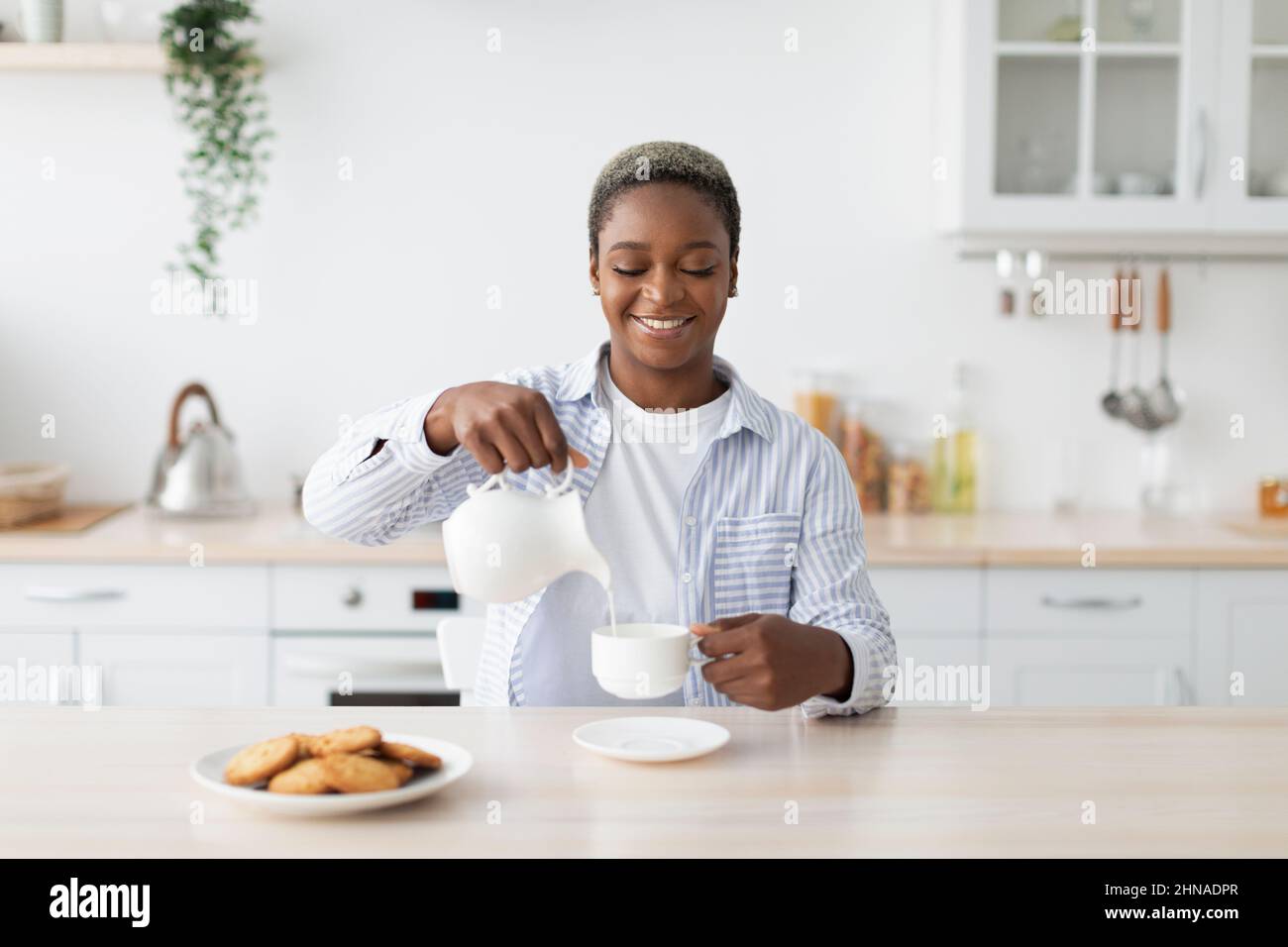 Smiling millennial african american woman in casual pouring milk at coffee at table minimalist kitchen interior Stock Photo