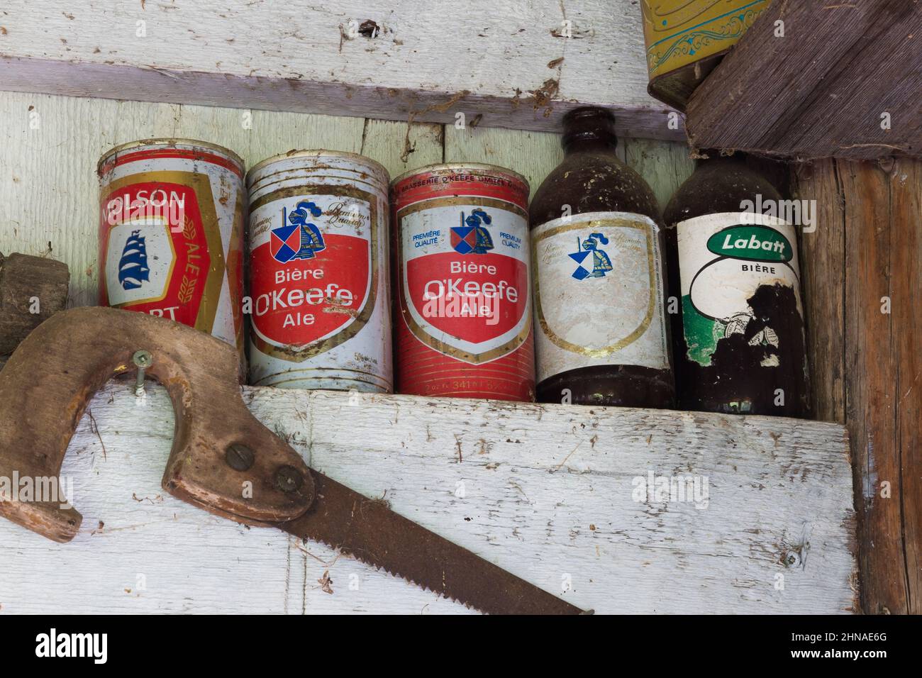 Old saw, beer cans and bottles on shelf inside old rustic wood plank cabin, Quebec. Stock Photo