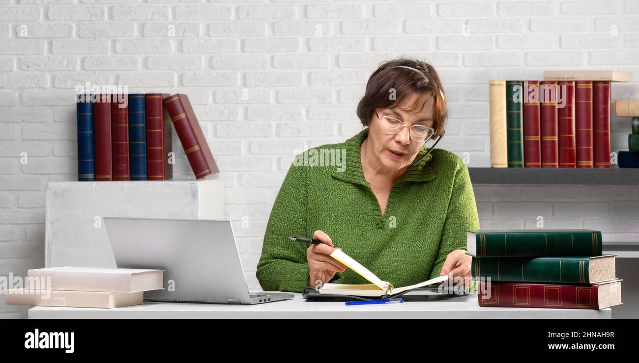A senior adult woman is working in front of a laptop monitor in the office - she is dictating notes from her notebook during an online call. Photo for Stock Photo