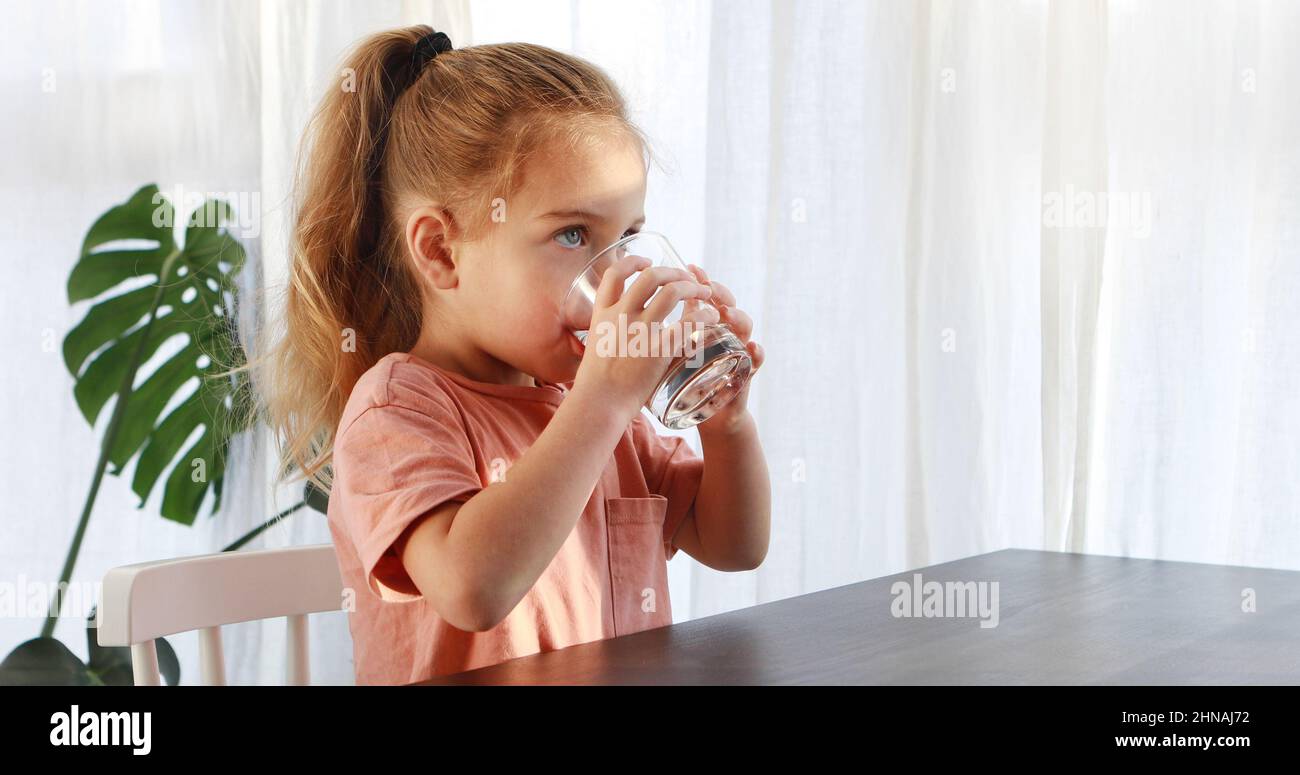 Little blonde girl in white dress drinks water from a glass indoors sunny day. Cute child is drinking a cup of water Stock Photo