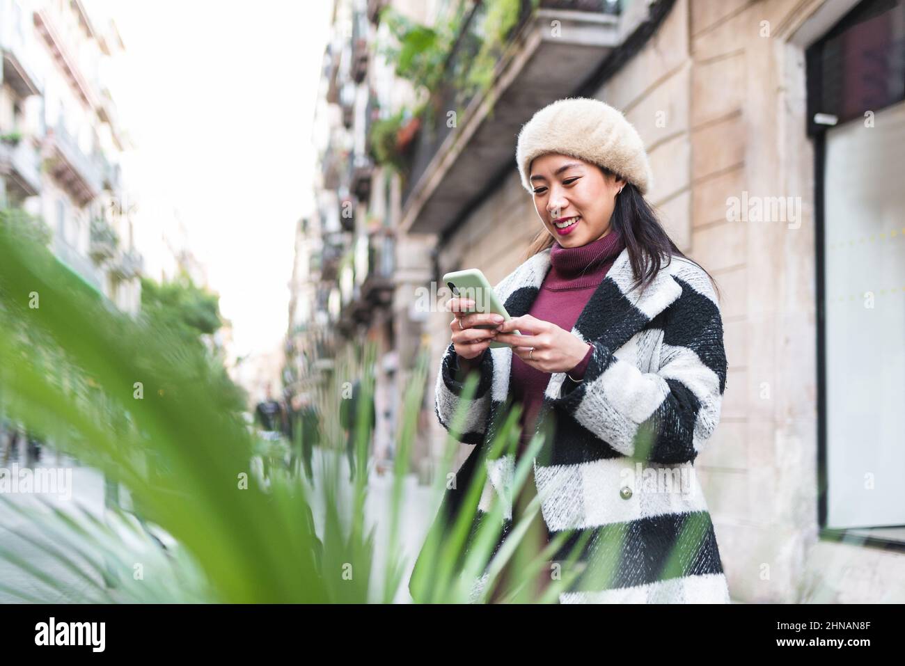 Positive Chinese female in outerwear text messaging on modern cellphone while standing on street near blurred green plants in city Stock Photo