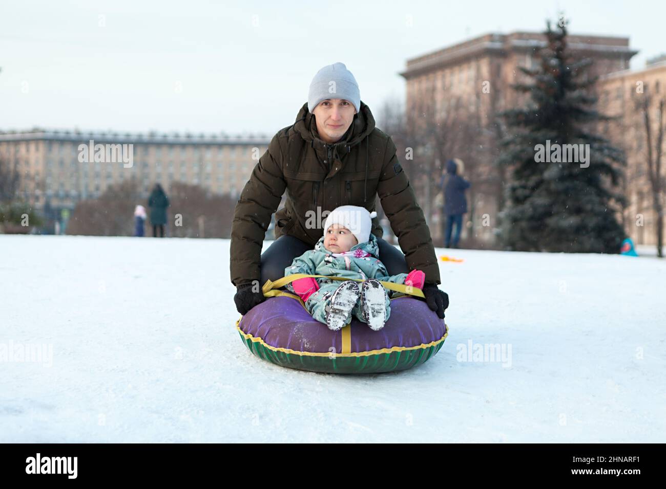 Father push her child from snowy slope, kid sitting in an inflatable toboggan, winter season is in Russian city Stock Photo