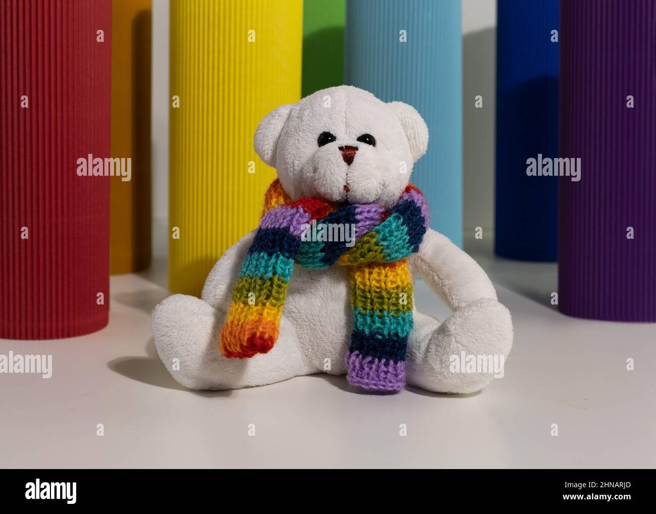A white teddy bear toy with a knitted scarf in rainbow colors sits on a ...