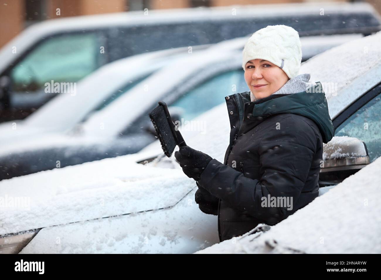 Female driver removes snow from the front of snowy car, woman looking at camera Stock Photo