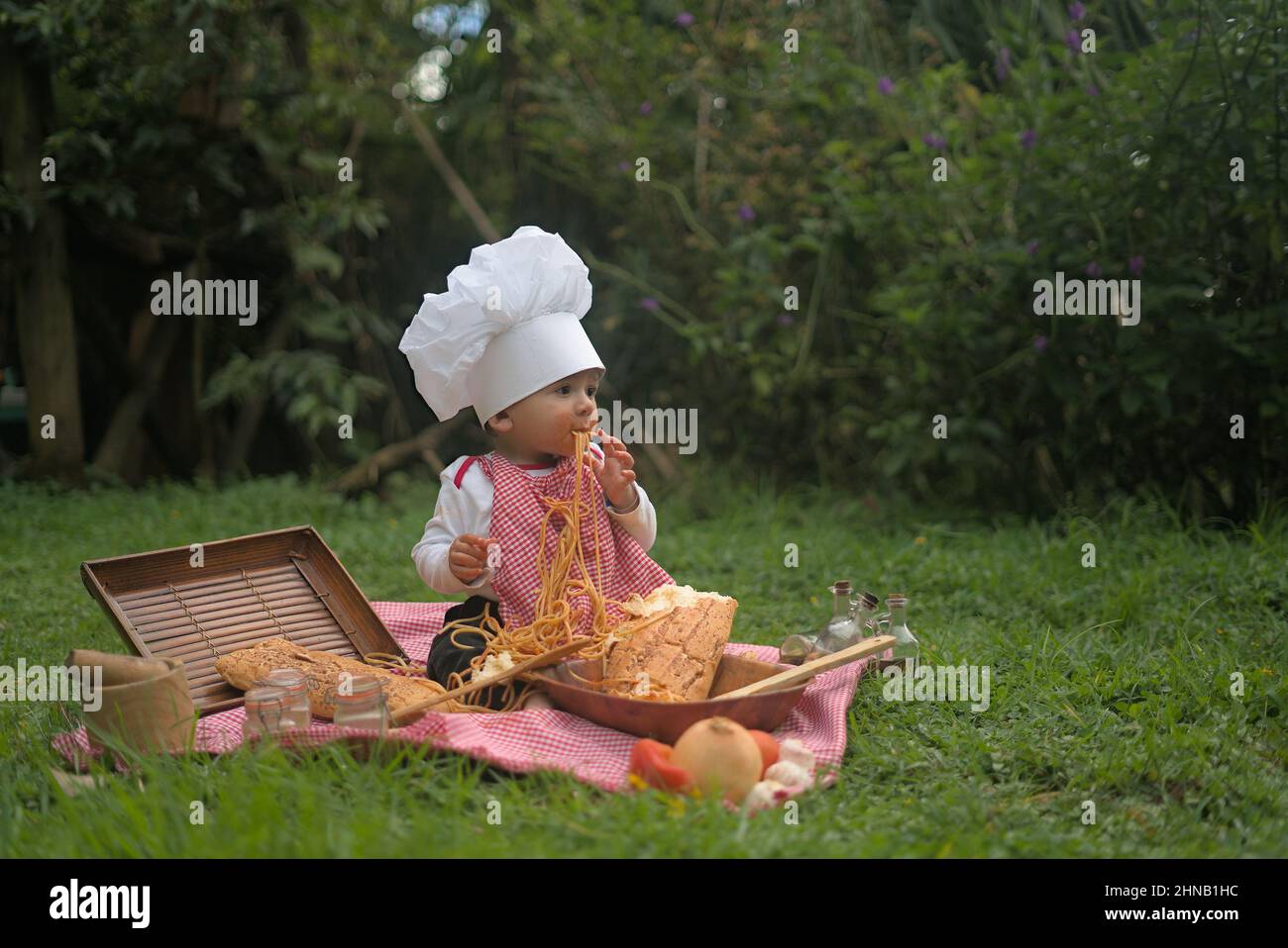 Cute Caucasian kid eating pasta on the grass in a chef uniform Stock Photo