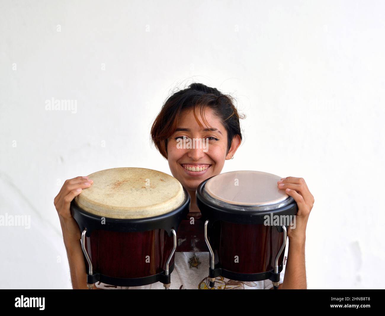 Cheerful young pretty Mexican female bongo player (percussionist) smiles with her pair of bongos (bongo drums) and looks at the viewer. Stock Photo