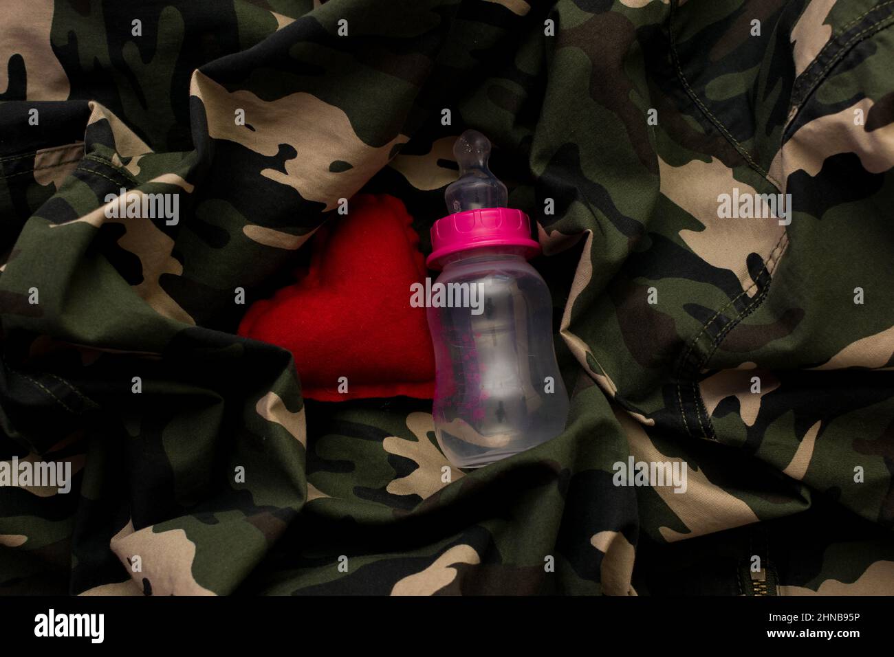 Red heart and baby bottle on military uniform, top view. Love and war concept Stock Photo