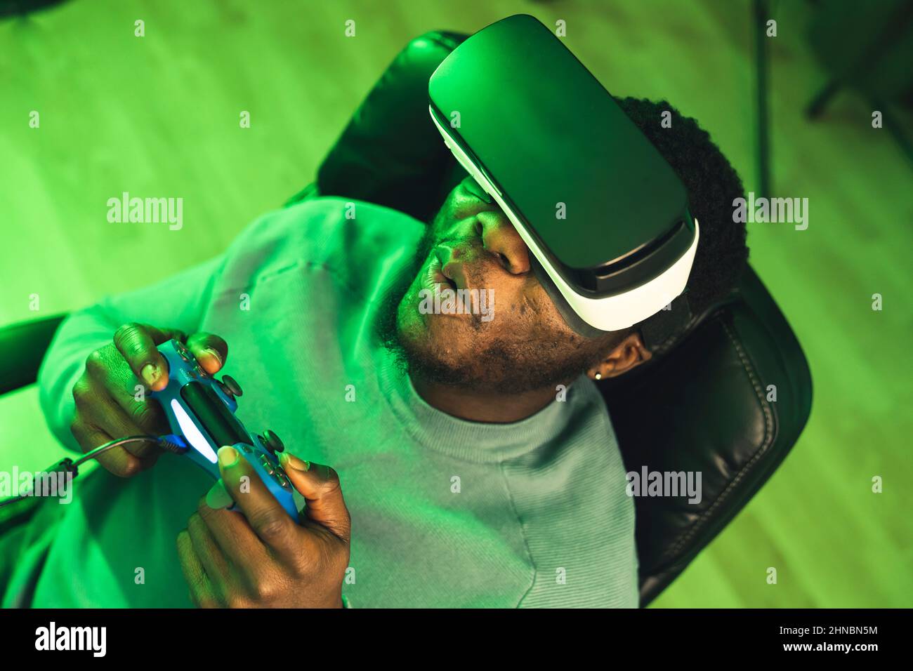Suprised impressed afroamerican black man playing the newest game on vr headset with a gaming pad sitting on black office chair surrounded by green ambient light - overhead portrait. High quality photo Stock Photo