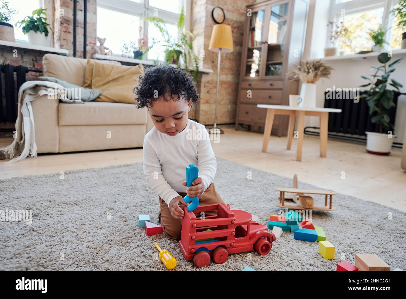 Serious little boy with curly hair sitting on carpet and playing car mechanic at home Stock Photo