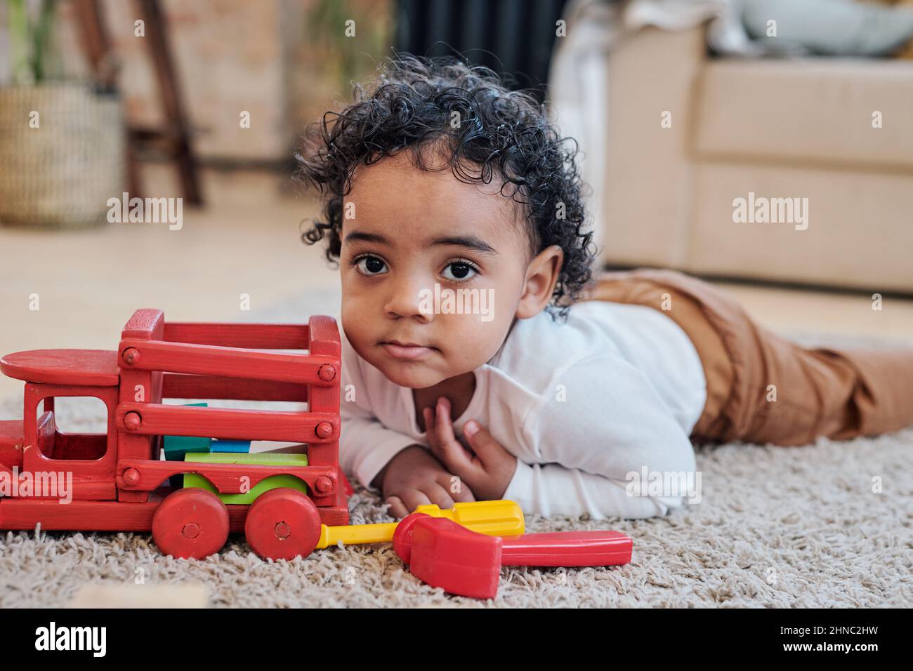 Portrait of serious curly-haired black boy lying near toy car on carpet in living room Stock Photo