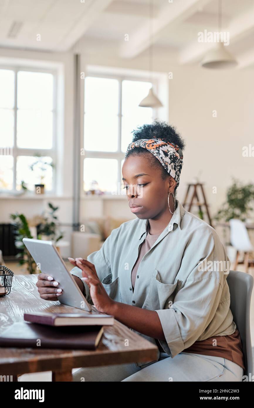 Serious young African woman in headscarf with abstract pattern sitting at desk and using tablet in cozy office Stock Photo