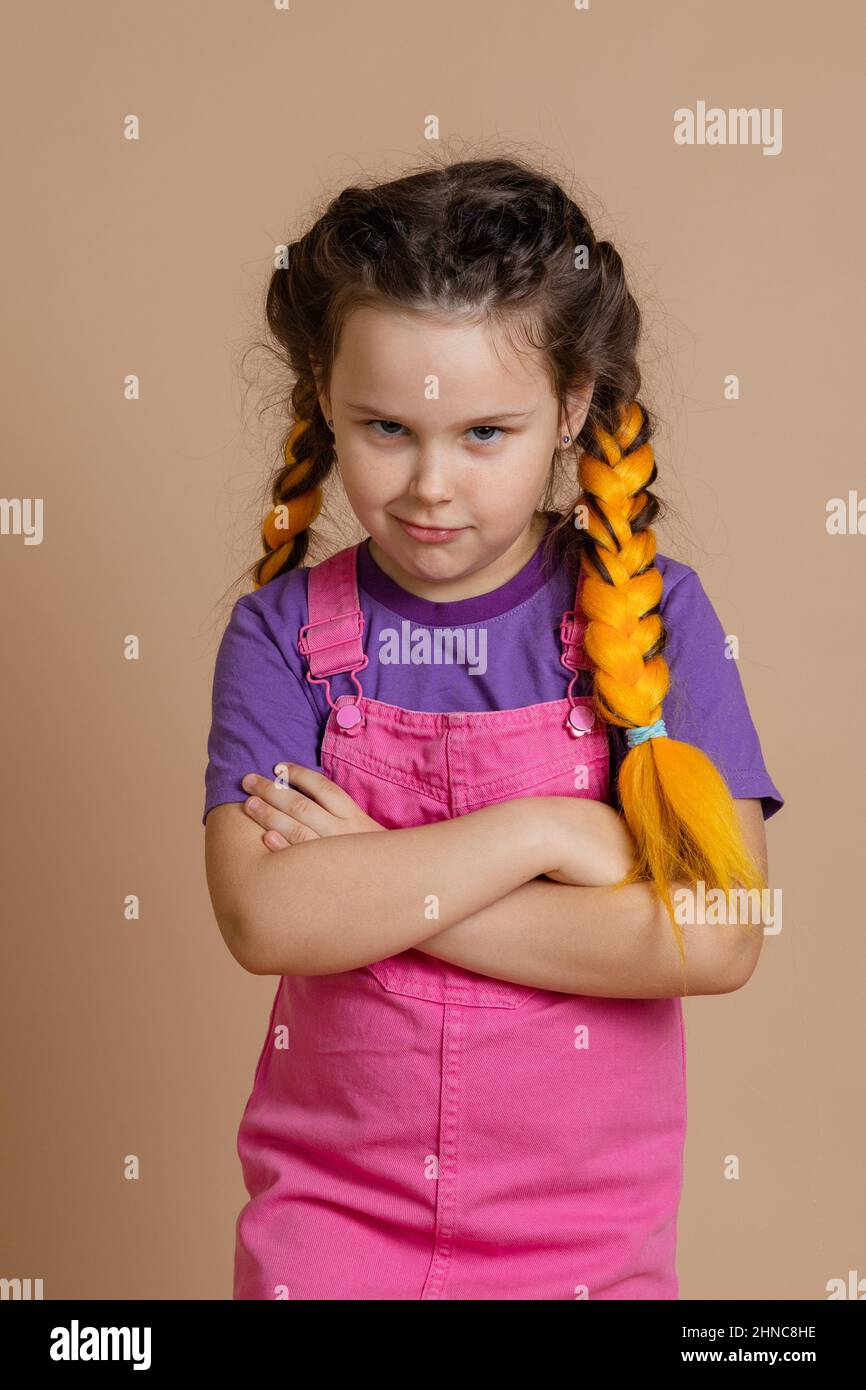 Offended small girl having yellow kanekalon braids looking at camera askance with folded arms wearing pink jumpsuit and purple t-shirt on beige Stock Photo