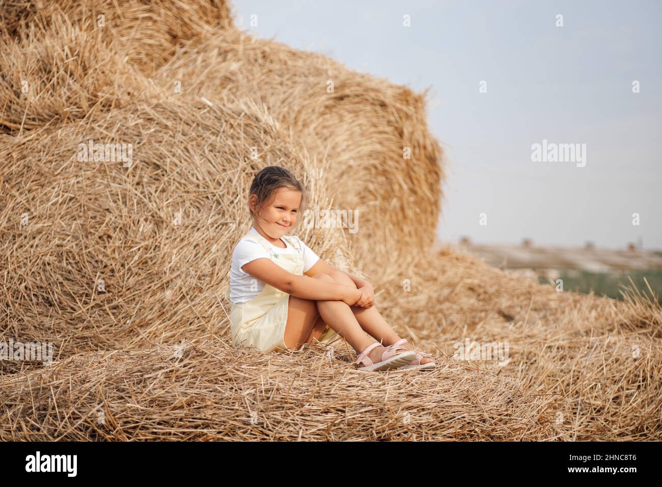 Small female kid with sly facial expression on haystack looking somewhere away slightly smiling. Time away from city in country field with tons of Stock Photo
