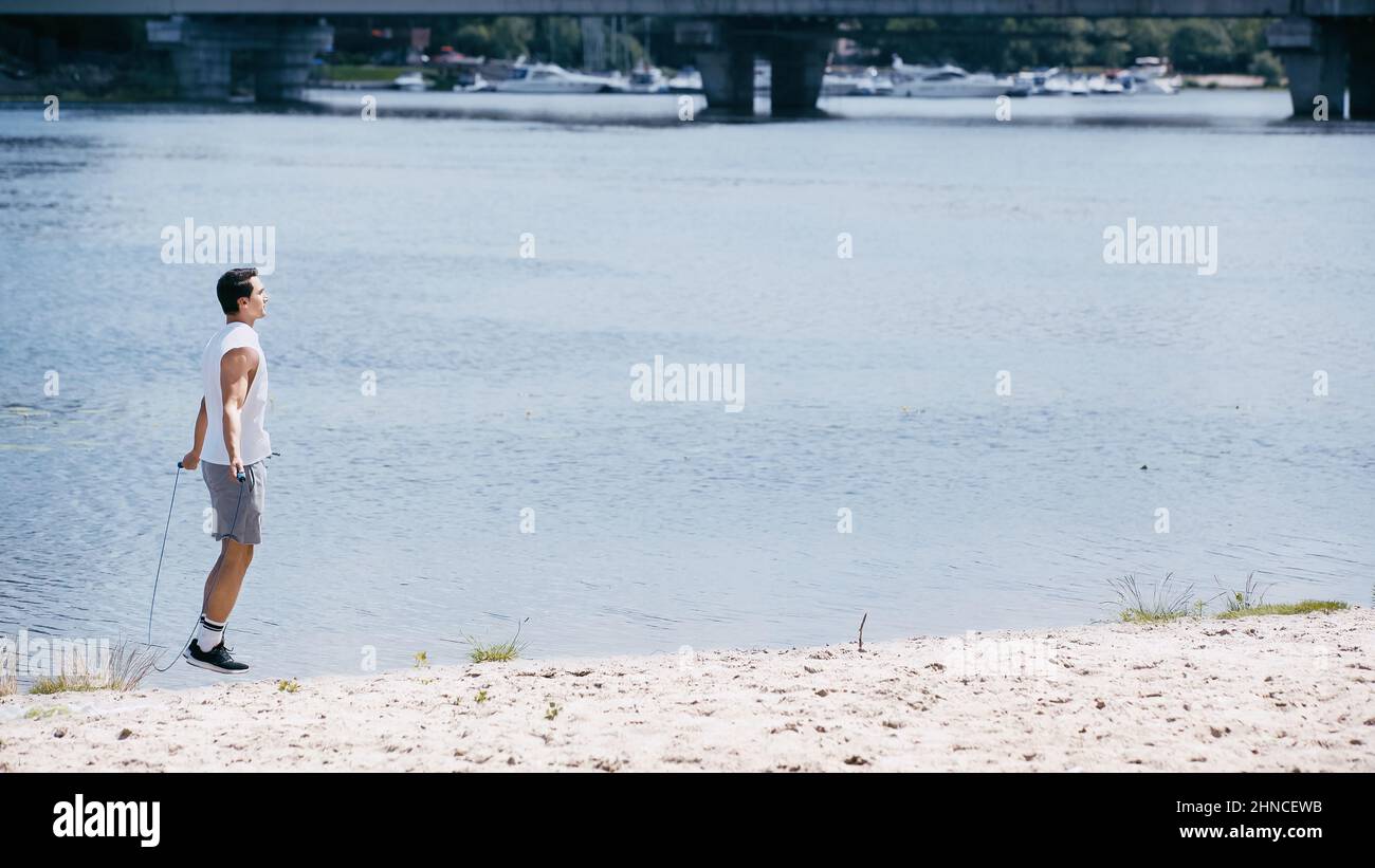 side view of young sportsman jumping with skipping rope on sand near