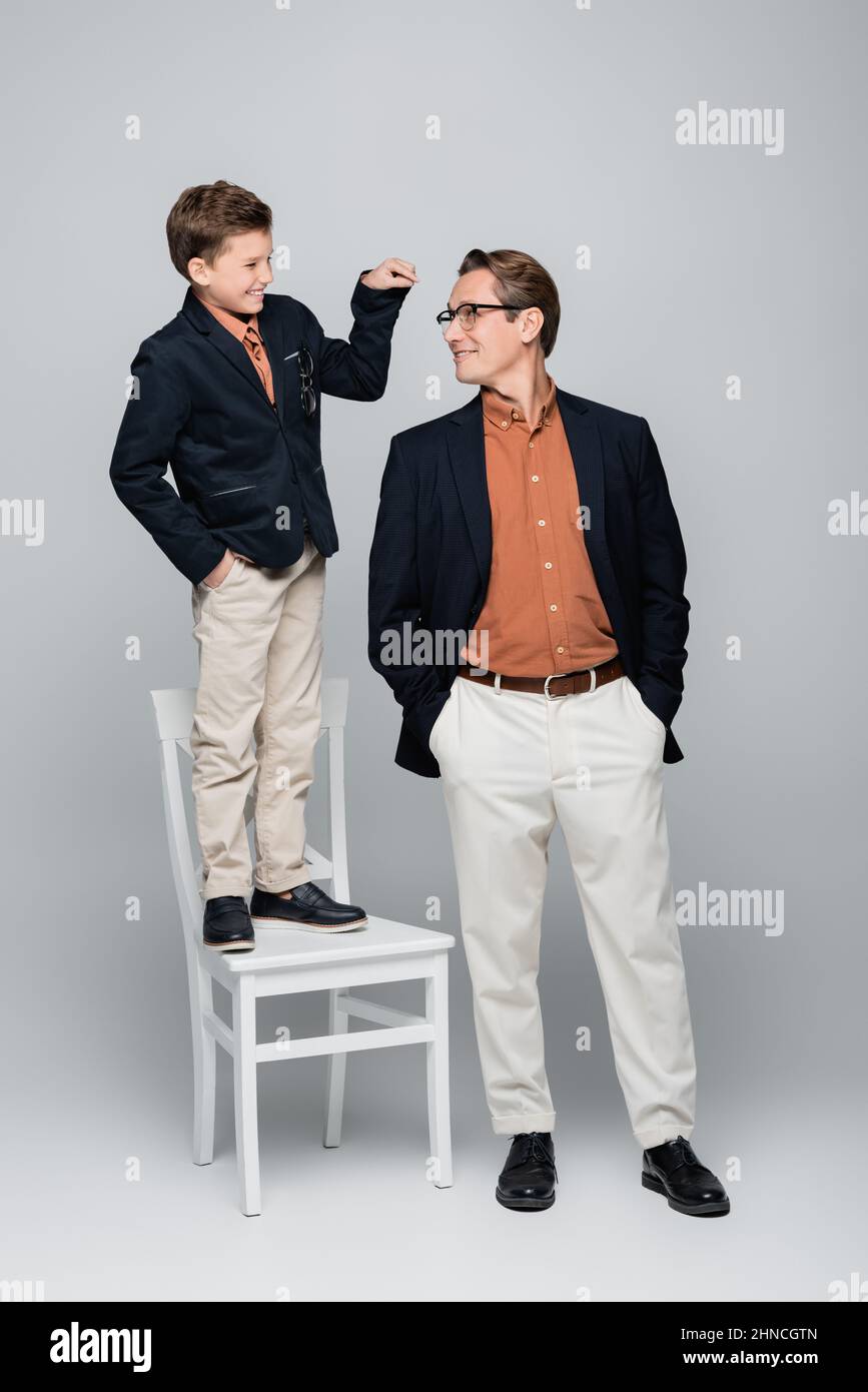 Smiling boy pointing at stylish dad while standing on chair on grey background Stock Photo