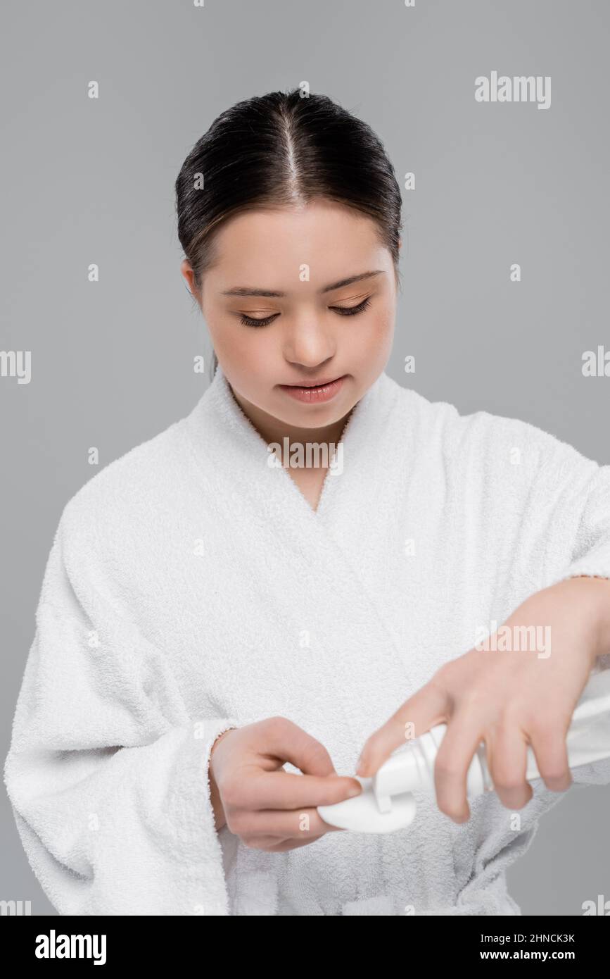 Young woman with down syndrome in bathrobe pouring cleansing foam on cotton pad isolated on grey Stock Photo