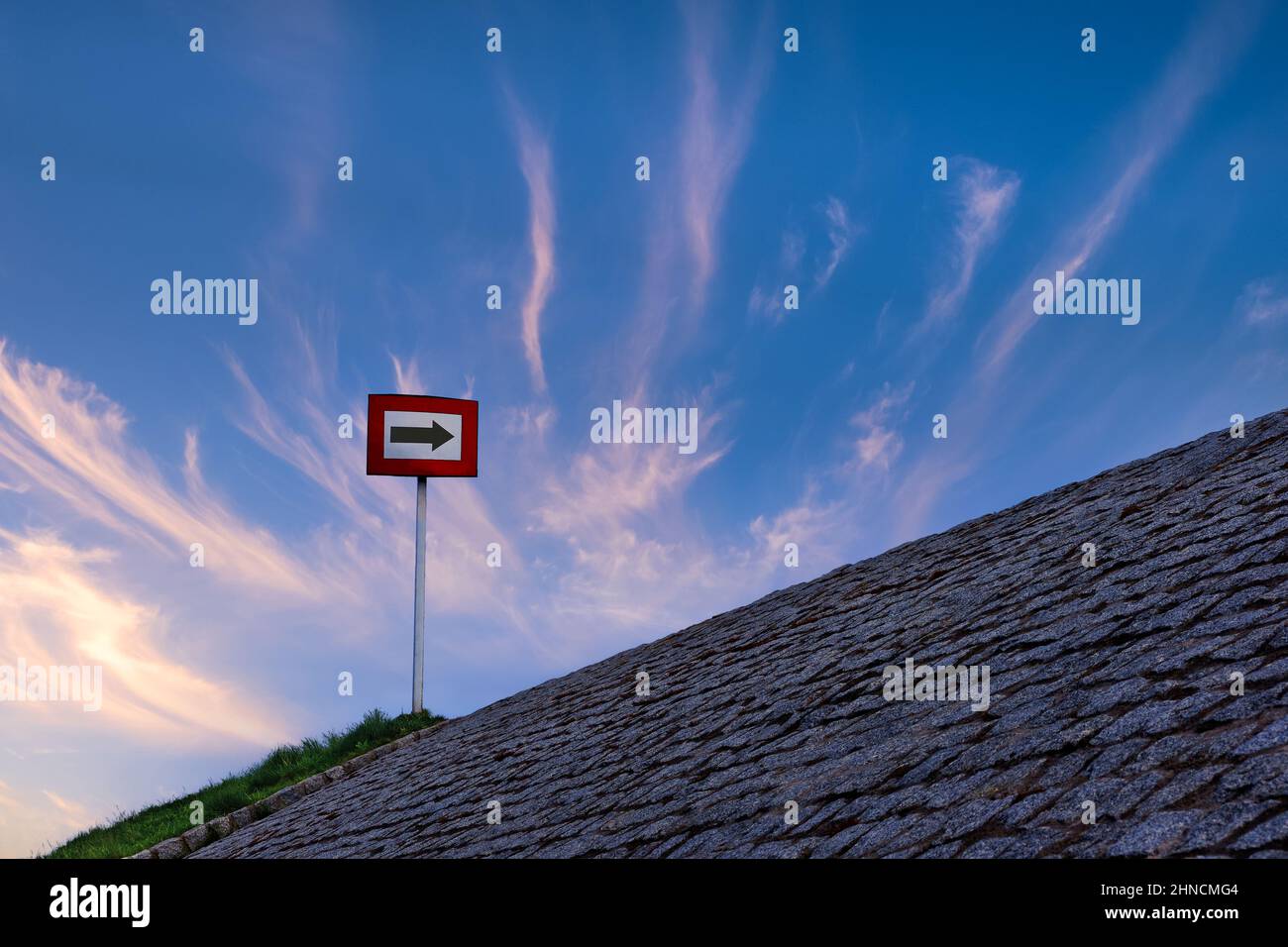 Black arrow on white background and in red frame standing atop of cobblestone paved hill at sunset. Blue sky with great clouds. Copy space, concept Stock Photo