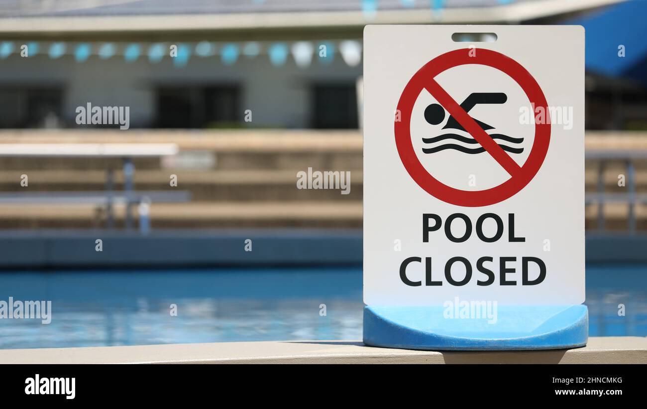 A bright white sign at a local outdoor pool facility saying pool is closed. No entry to water. Clear clean blue water at public baths and no swimming Stock Photo
