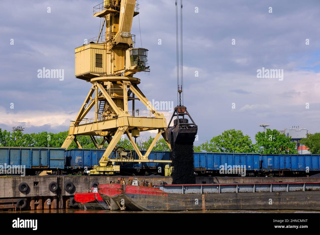 River port crane with clamshell or griper loading coal to river drag boats or barges moored by pier on cloudy day. Energy, minerals, environment  Stock Photo