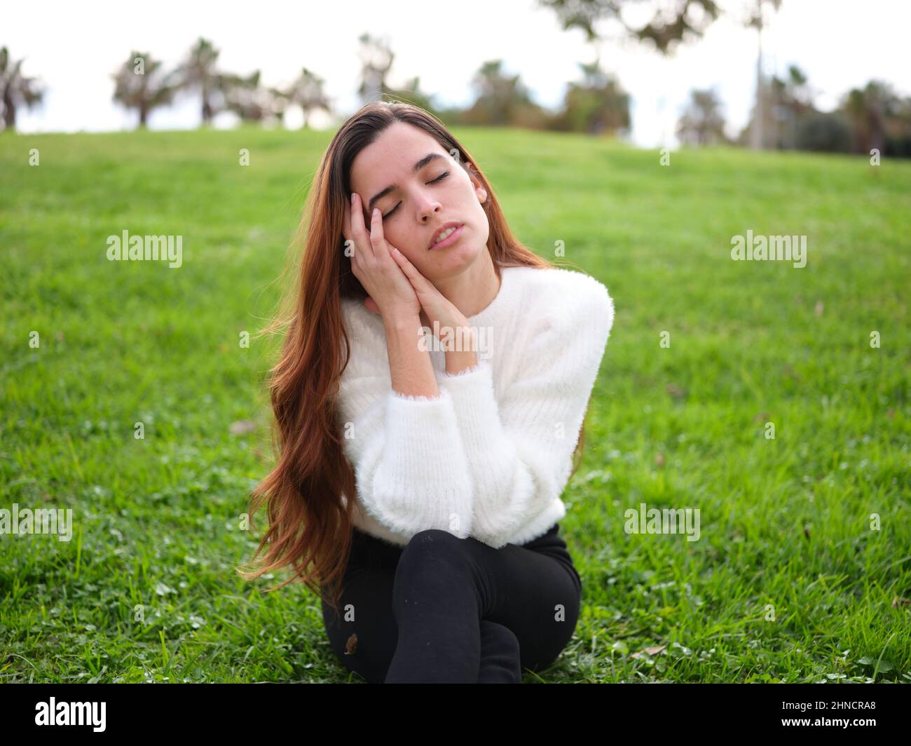 A young woman in the park sitting on the grass with her hands on her face and her eyes closed daydreaming. Stock Photo