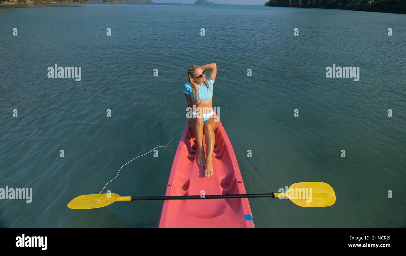 Pretty slim woman in elegant swimsuit lies in pink plastic kayak drifting on azure ocean at exotic resort upper view. Traveling to tropical countries. Girl is sailing on kayak in ocean, view above. Stock Photo