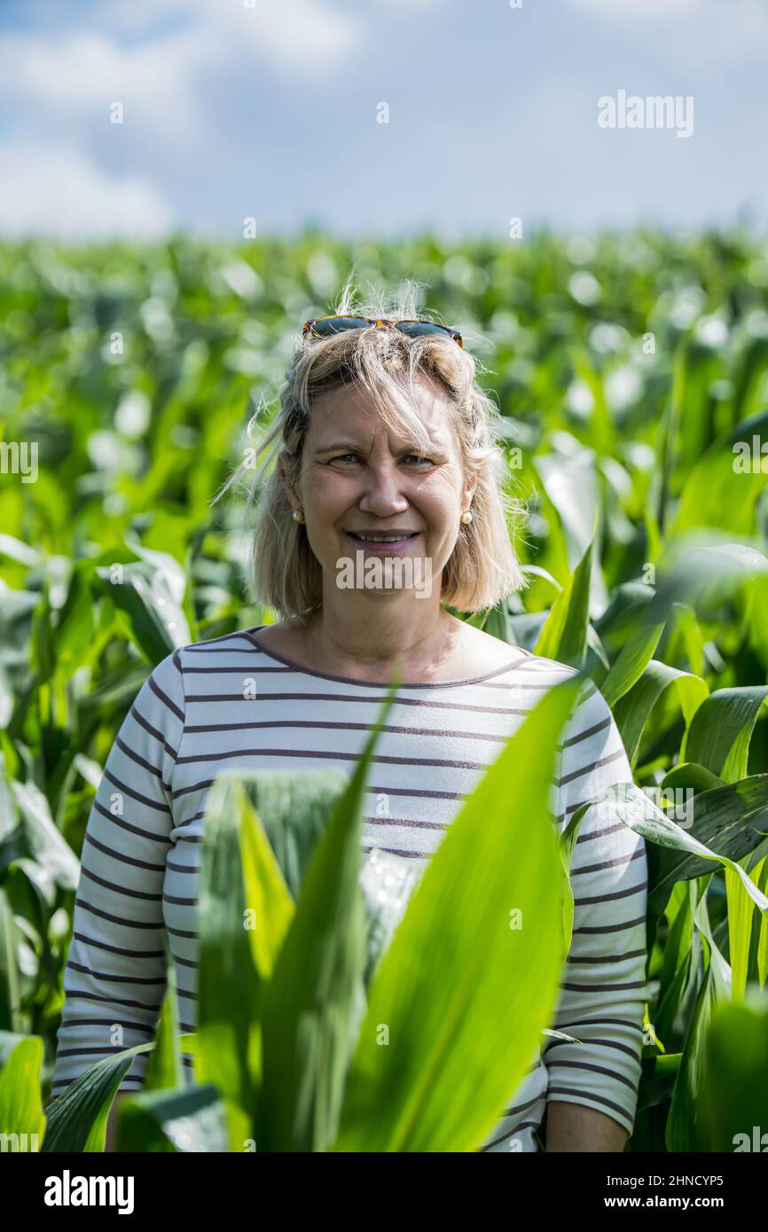 Glad middle aged female looking at camera with smile while standing in plantation with green plants on summer day in France Stock Photo