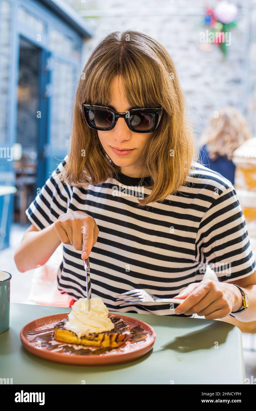 Smiling female in sunglasses eating delicious sweet waffles with ice cream while sitting at table on terrace on cafeteria on summer day Stock Photo