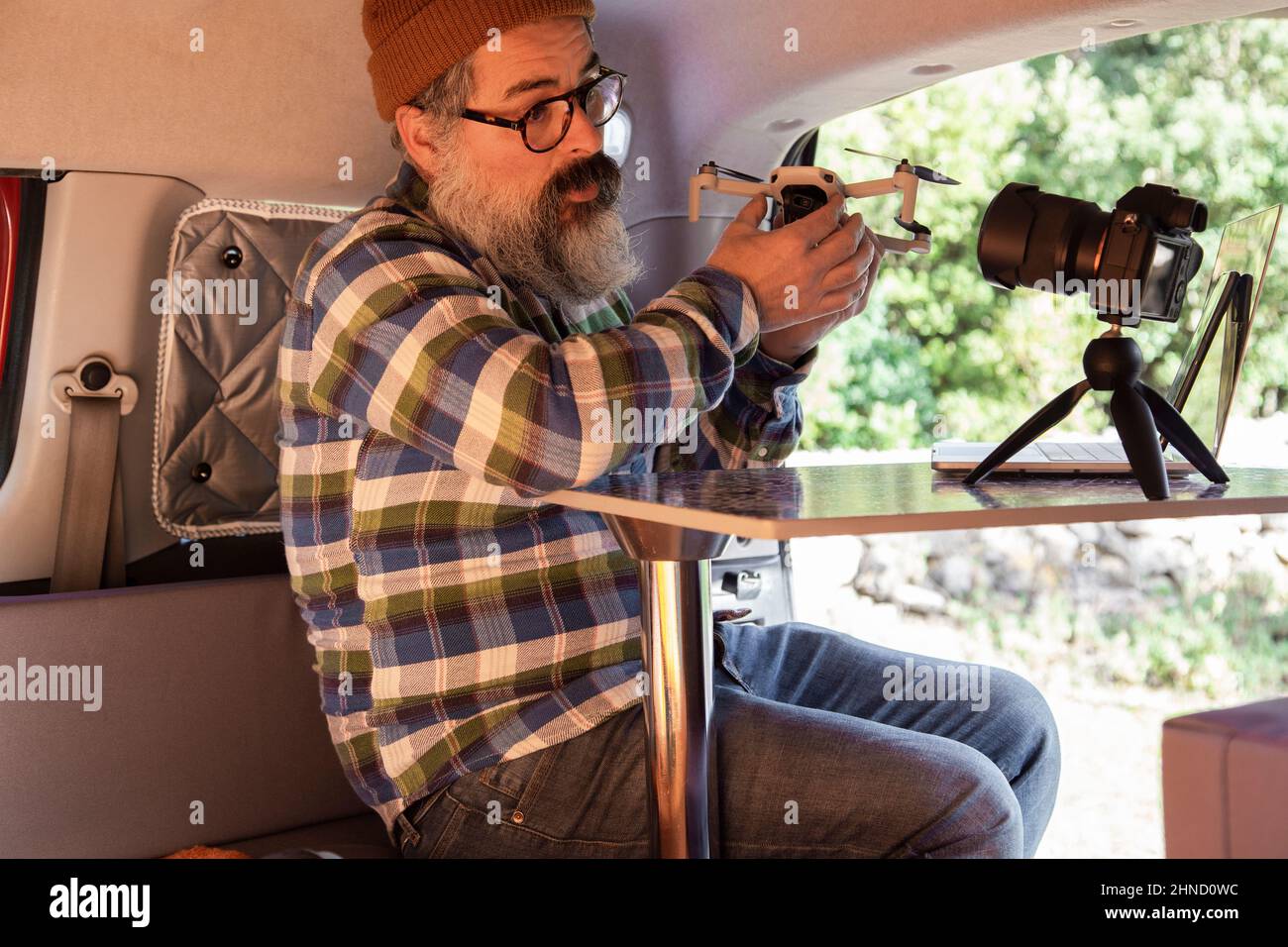 Mature bearded male blogger with modern drone recording video on camera while sitting at table in mini camper parked in countryside Stock Photo