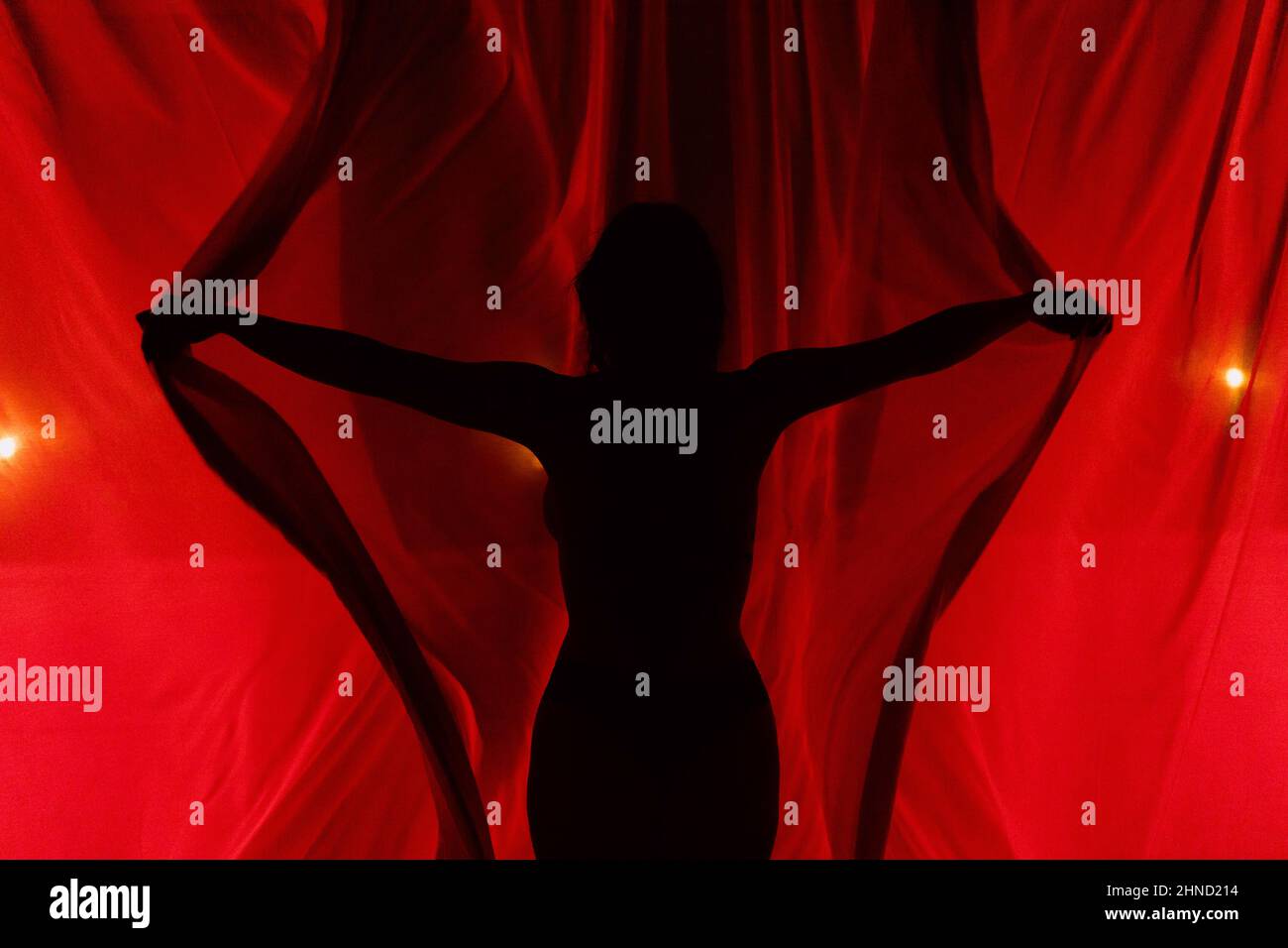 Silhouette of unrecognizable female opening light red curtains with spread arms while standing in murk room at dark night time Stock Photo
