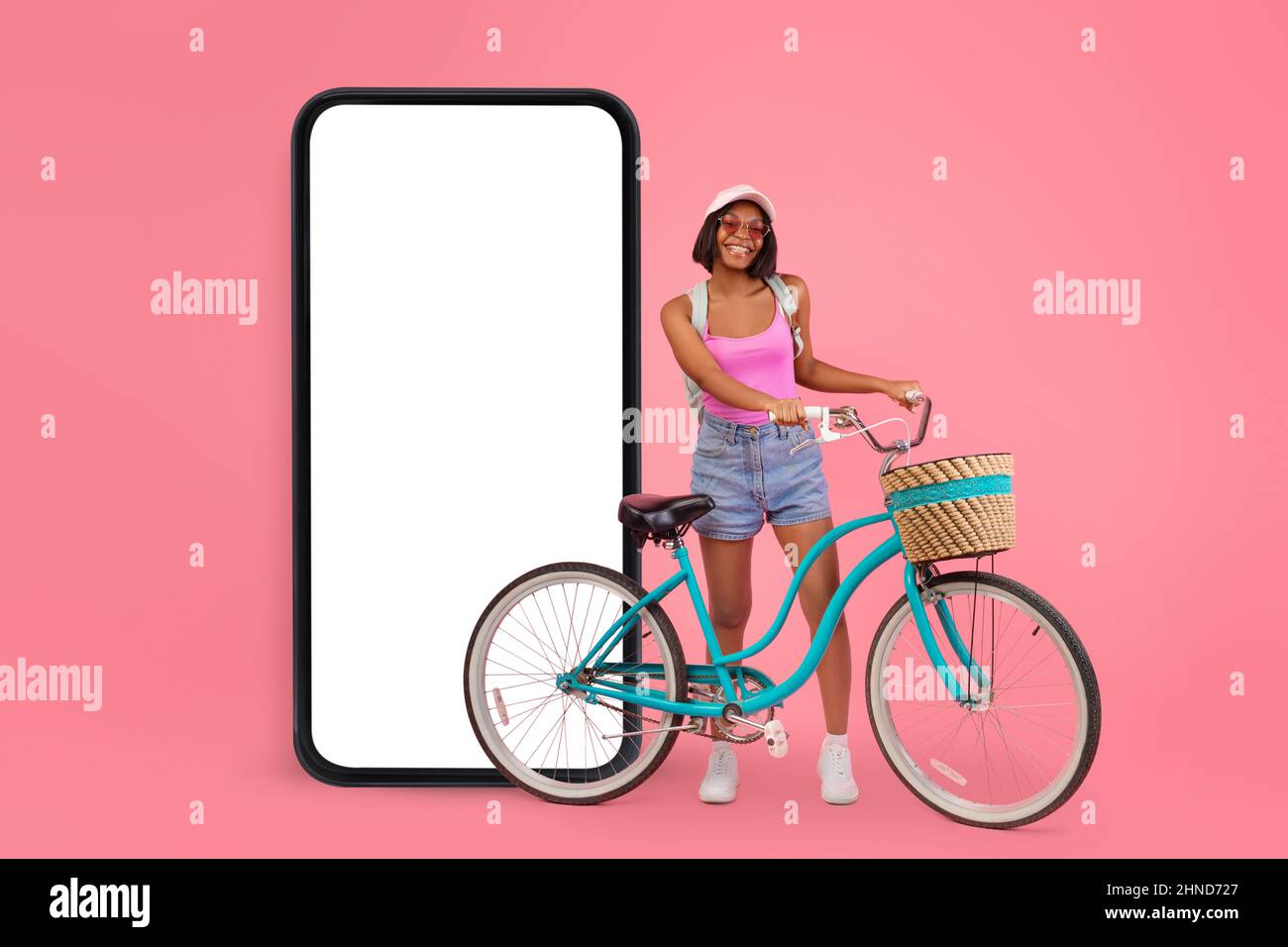 Mobile offer. Black lady riding her bike near huge smartphone with empty white screen over pink studio background Stock Photo
