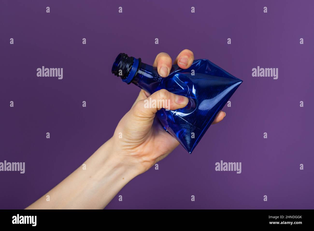 Woman's hand holds a crumpled blue plastic bottle on a purple background Stock Photo