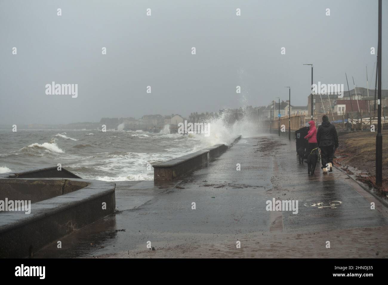 Prestwick, Scotland, UK; 16th February 2022. A High Tide ahead of Storm Dudley brings waves crashing up onto the Promenade at Prestwick Seafront, South Ayrshire.   Two women are walking their dogs along the promenade. Liz Leyden/Alamy Live News Stock Photo
