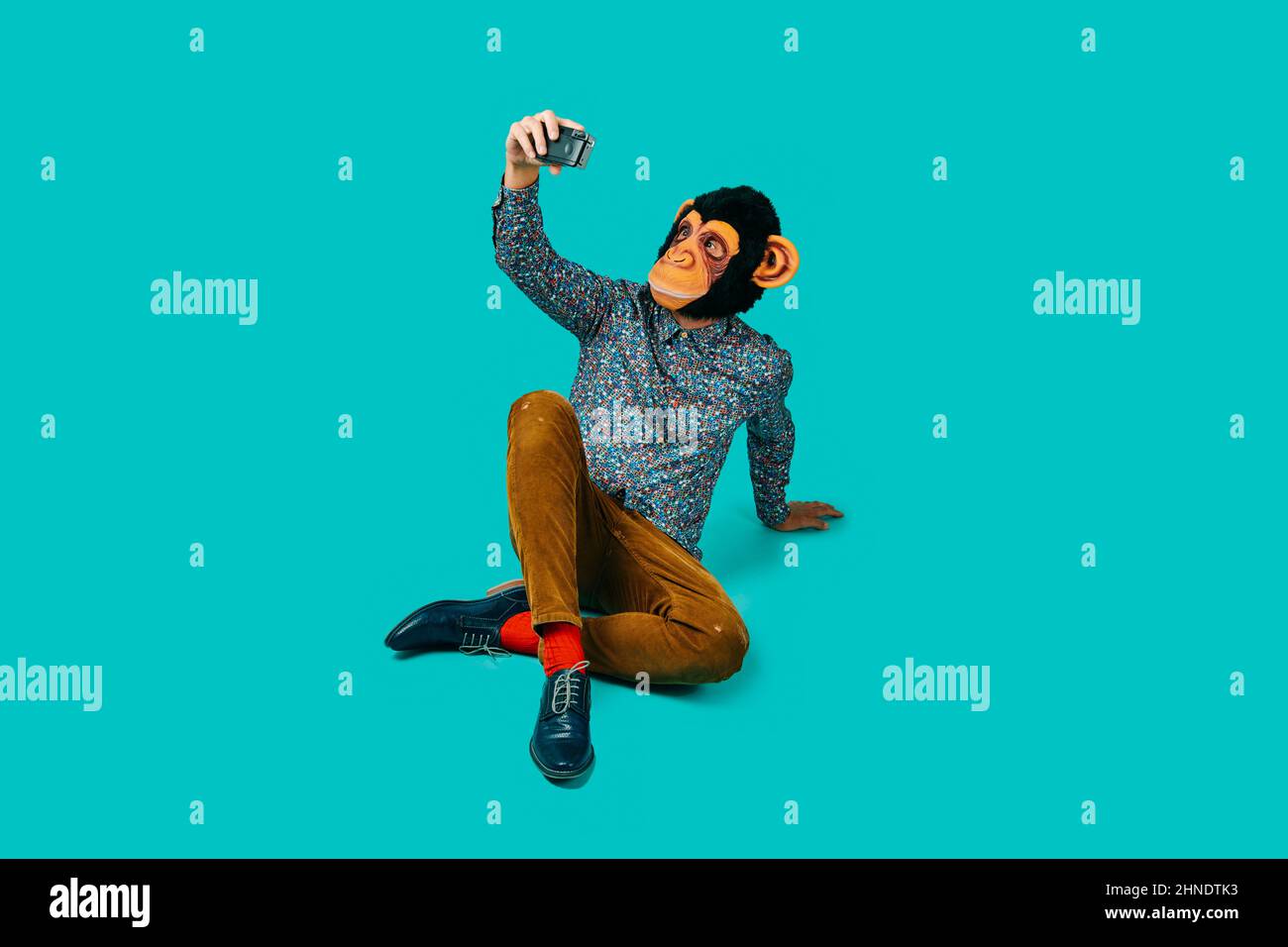 man, wearing a monkey mask and colorful clothes, taking a selfie with a retro film camera, sitting on a blue background with some blank space around h Stock Photo