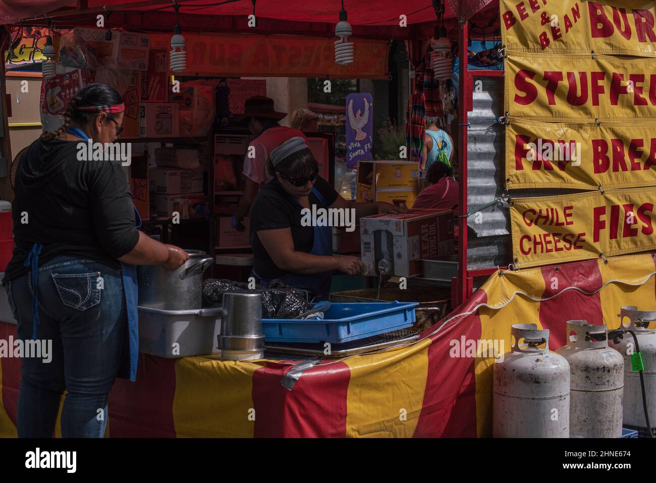 Two women with black hair work in a food booth, one pouring cooking oil for fried foods. A sign advertises fry bread and chile cheese fries. Stock Photo