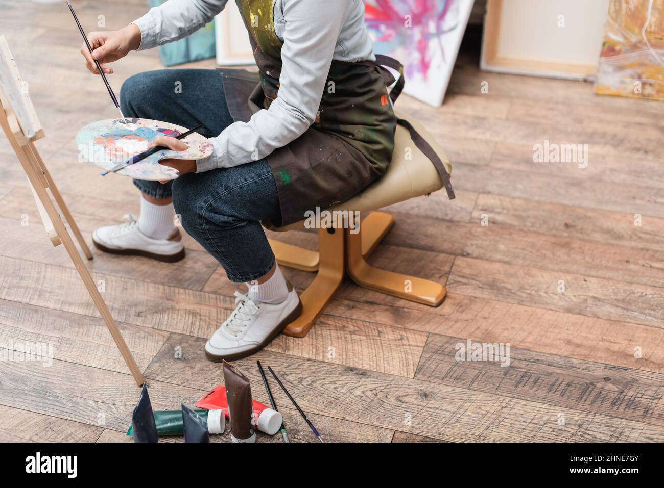 partial view of woman with palette and paintbrushes sitting on chair near easel Stock Photo