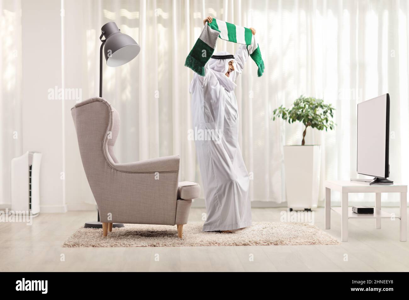 Full length profile shot of an arab man in a thobe with a scarf cheering in front of tv at home Stock Photo