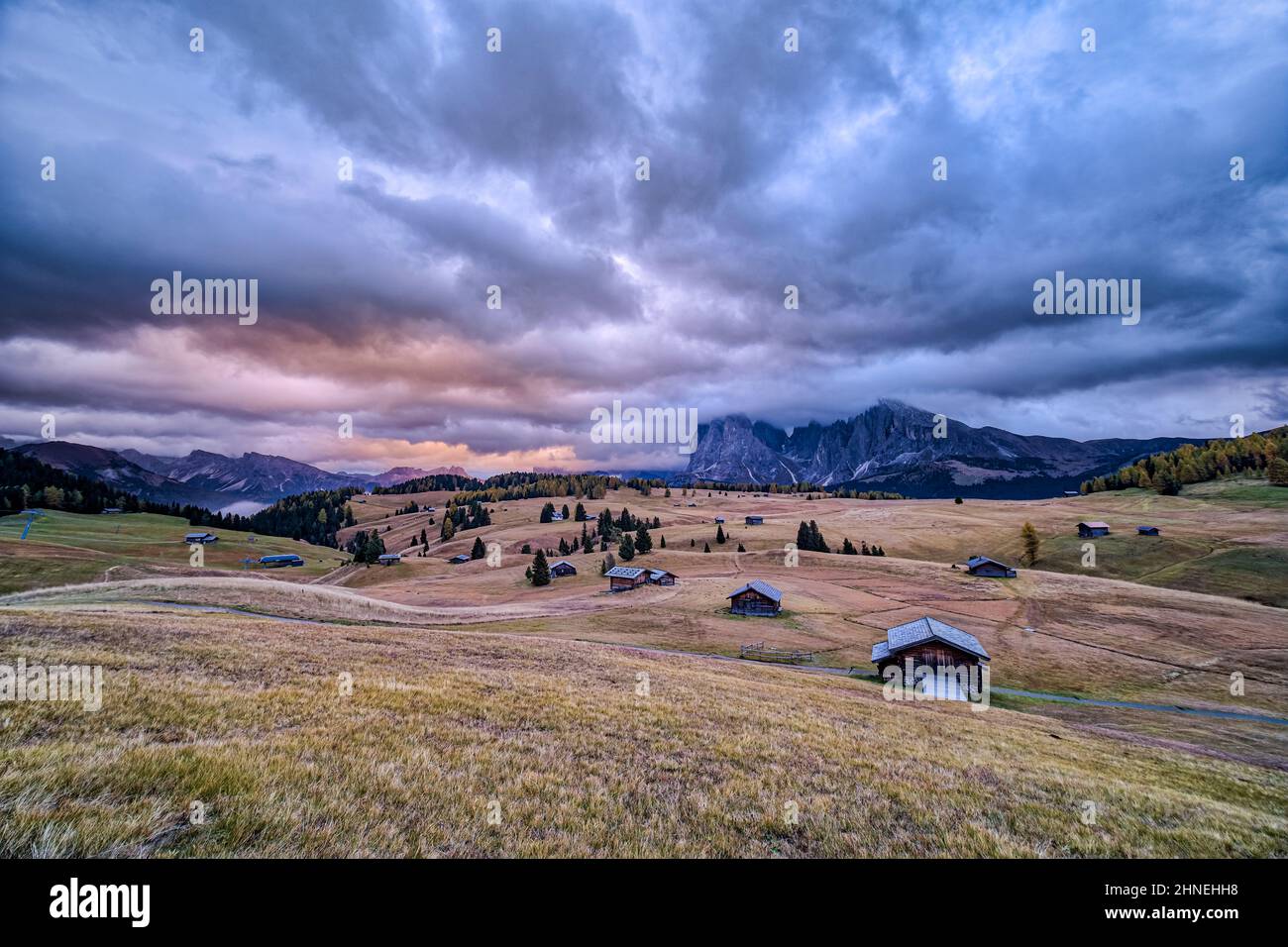 Hilly agricultural countryside with wooden huts at Seiser Alm, the mountains Langkofel (left) and Plattkofel (right) in the distance, at sunrise. Stock Photo