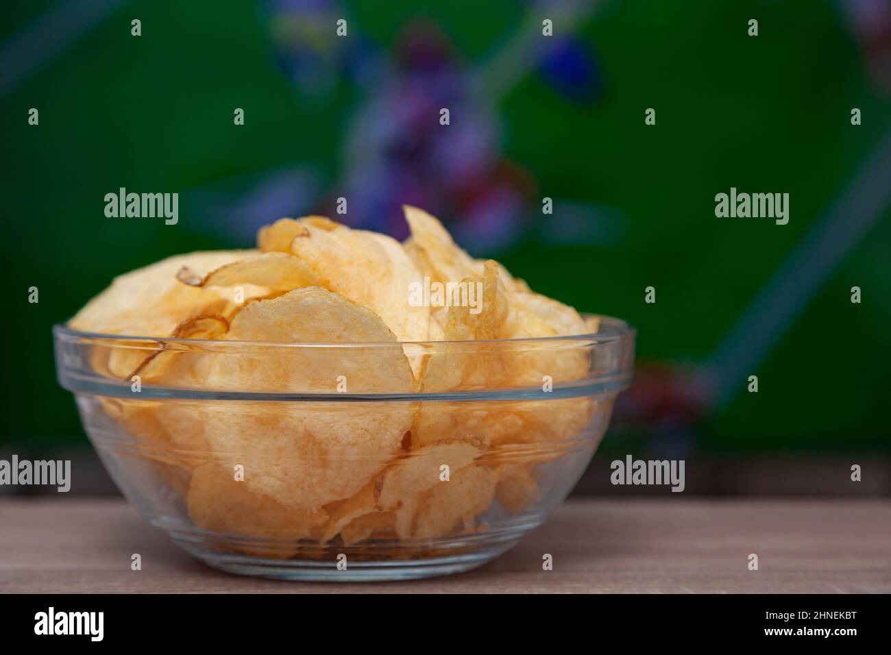 A clear glass bowl filled with chips on a wooden table. In the background, out of focus, is a television with a sports broadcast. Stock Photo