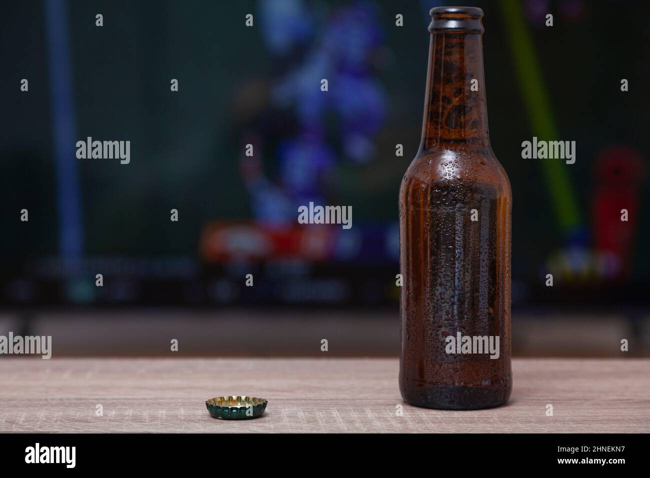 A bottle of fresh beer and a bottle cap on a wooden table. In the background, out of focus, there is a television with a sports broadcast. Stock Photo
