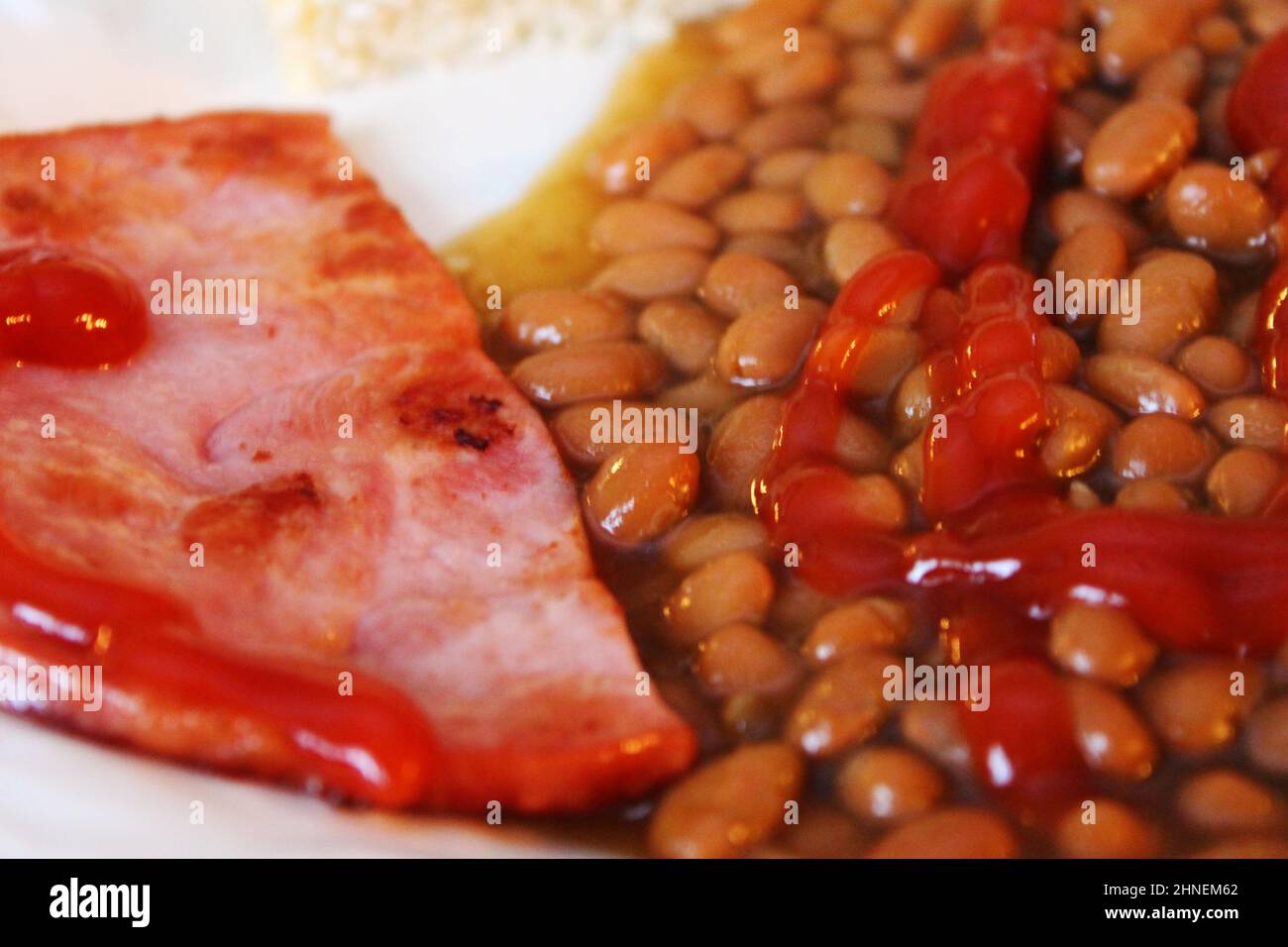 Close-up of a plate of baked beans, with fried ham, a slice of bread, and ketchup. Stock Photo