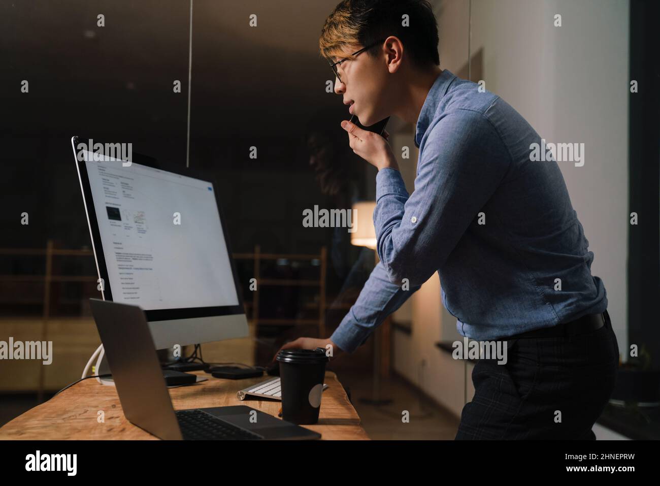 Focused asian guy talking on cellphone while working with computers at office Stock Photo