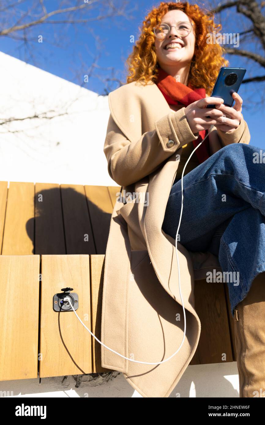 Young redhead woman charging her smartphone on a smart bench on a warm spring day Stock Photo