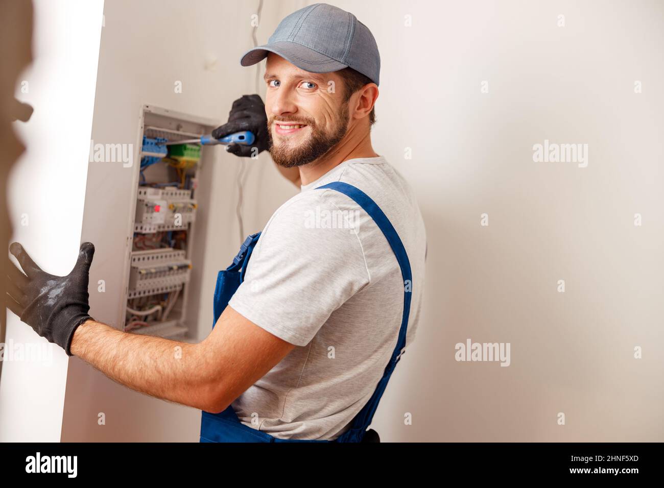 Electrical technician in uniform smiling at camera, using screwdriver while checking fuses in a switchboard on the wall Stock Photo