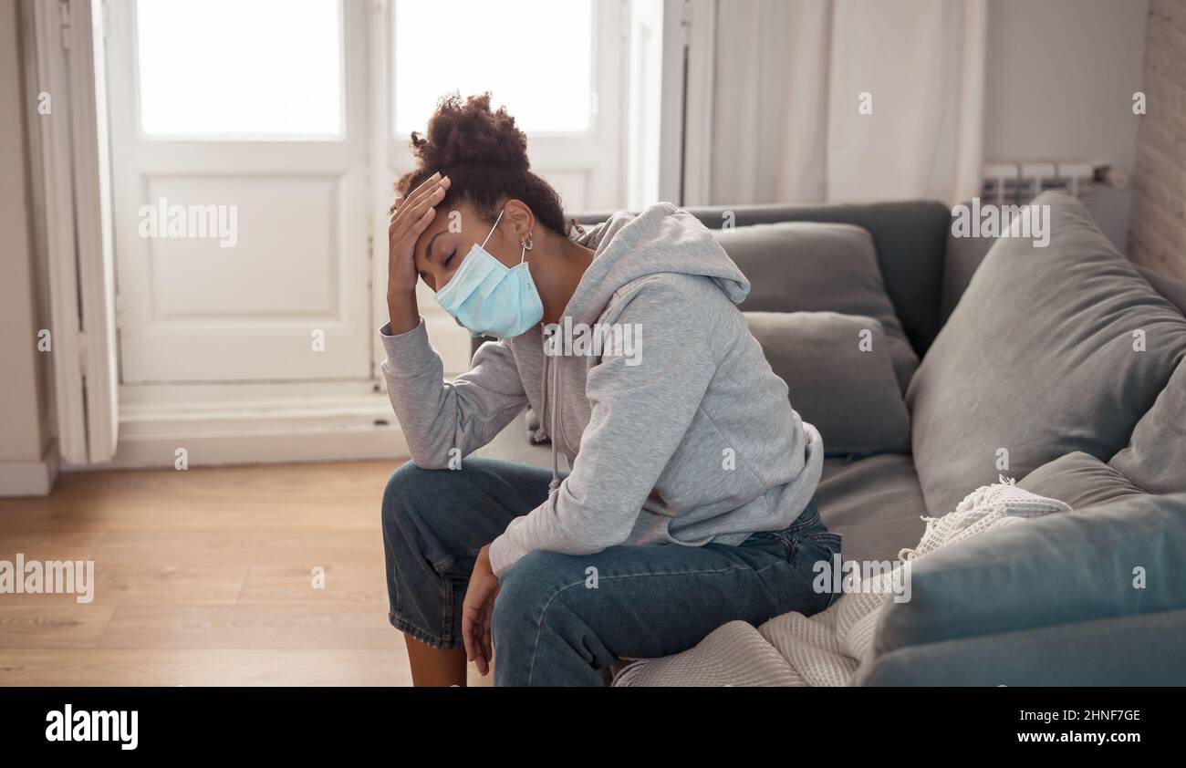 Young afro Latina young woman with a protective face mask on the sofa feeling ill, exhausted and sad. Depressed African American teen tired of stress, tension Stock Photo