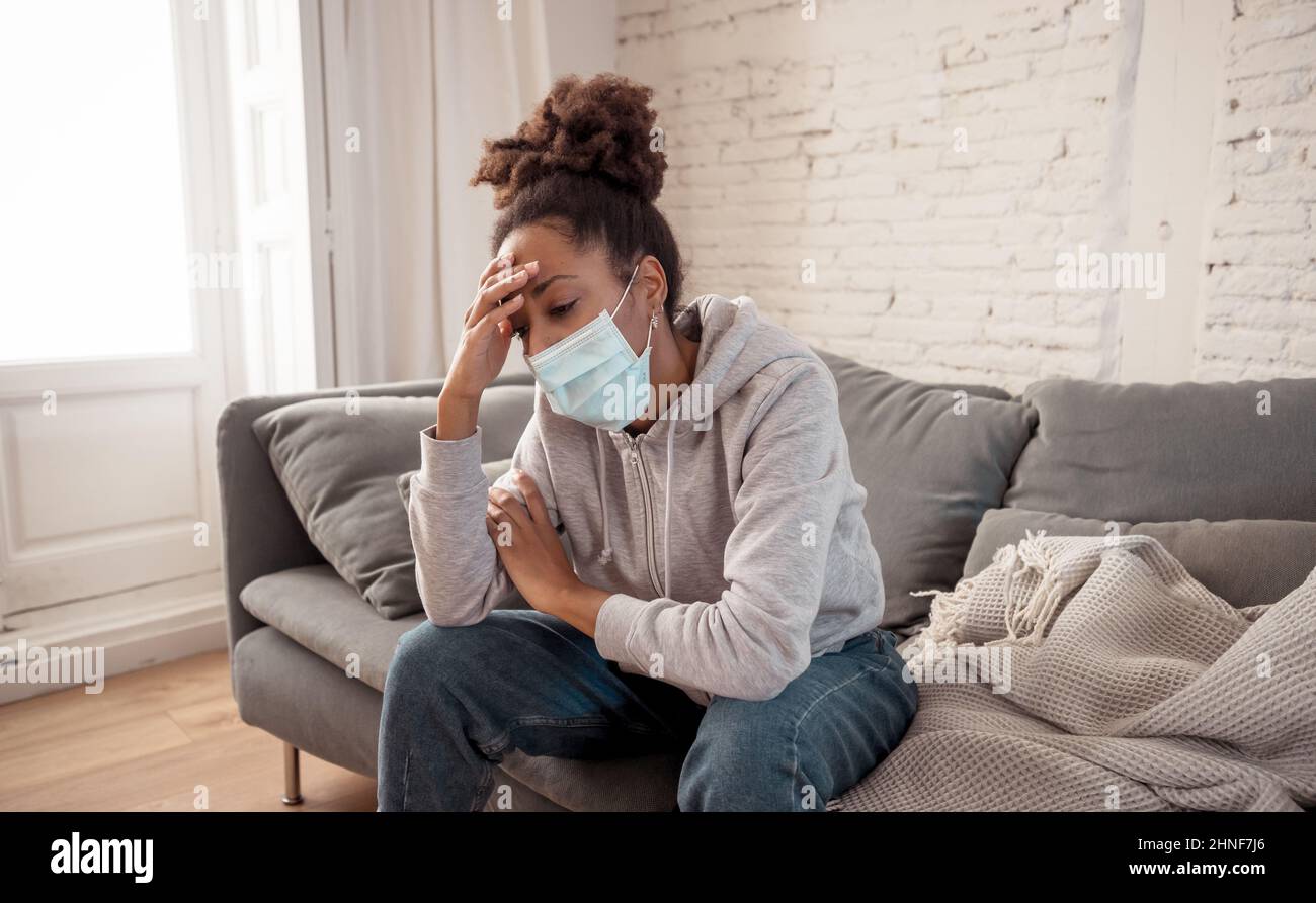 Young afro Latina young woman with a protective face mask on the sofa feeling ill, exhausted and sad. Depressed African American teen tired of stress, tension Stock Photo