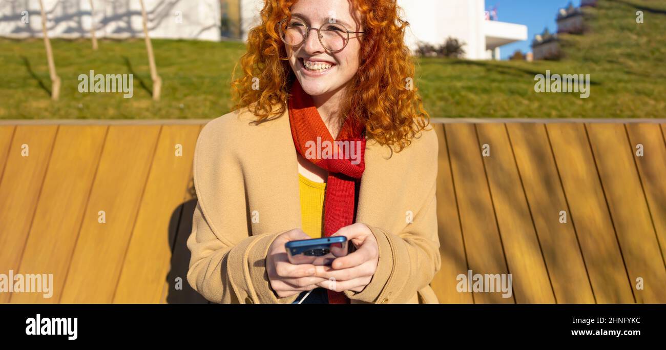 Young woman smiling while using her smartphone on a bench Stock Photo